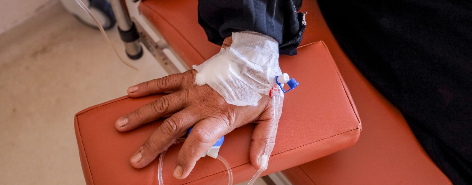 Close-up of a patient's hand with an IV line taped in place and a pulse monitor clipped to a finger, resting on a hospital blanket — the kind of outpatient infusion care Medicare Part B covers for Pitkin County cancer patients in 2026