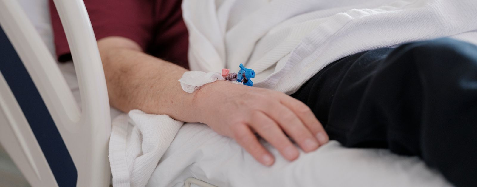 An older patient's weathered hand, with a pulse monitor clipped to a finger, held by a caregiver over a tan blanket in a hospital bed — the kind of ongoing kidney care Medicare covers for Delta County dialysis patients in 2026