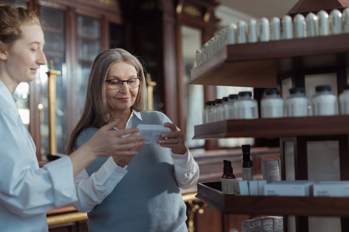 An older woman with glasses reading a medication box with a pharmacist beside the shelves, the kind of pharmacy visit where a Medicare Part D plan's preferred or standard status decides the copay in Glenwood Springs, Rifle, Carbondale and Parachute