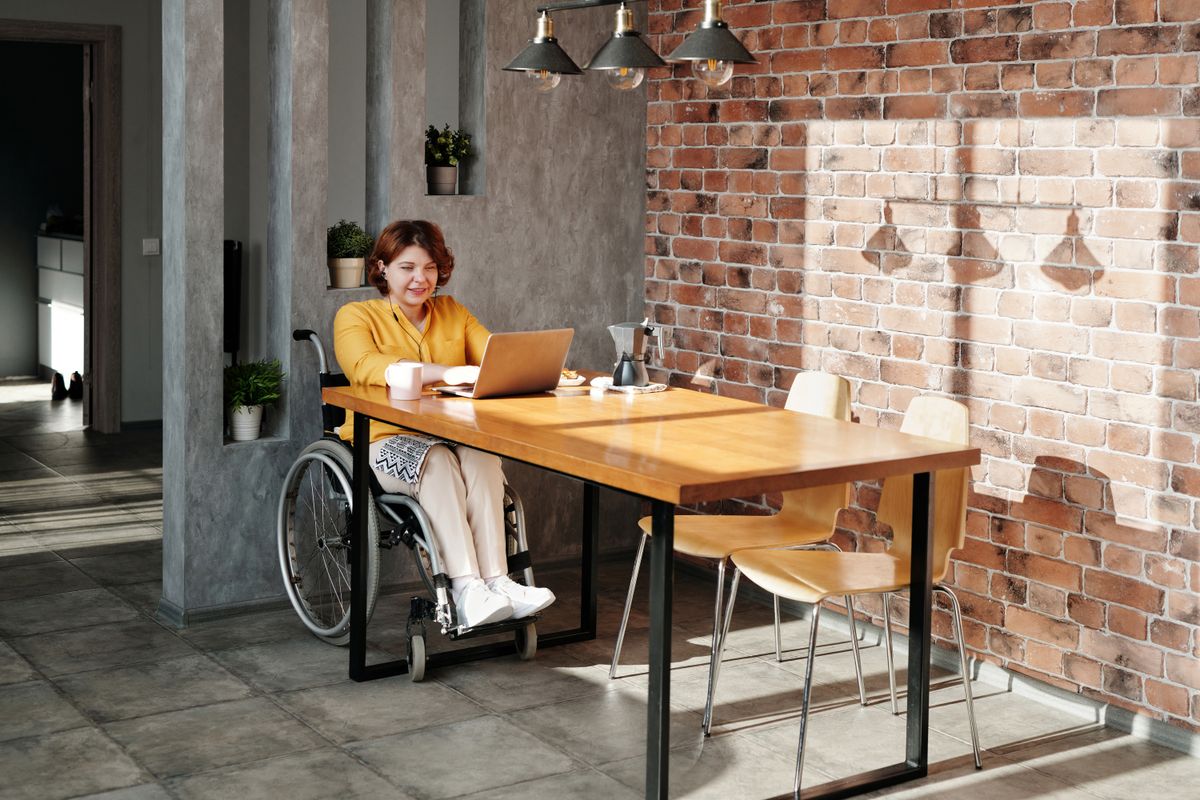 A woman in her forties seated in a wheelchair at a sunlit kitchen table, working on a laptop with a coffee cup beside her, the way a Roaring Fork Valley resident on Social Security disability might review her Medicare paperwork at home