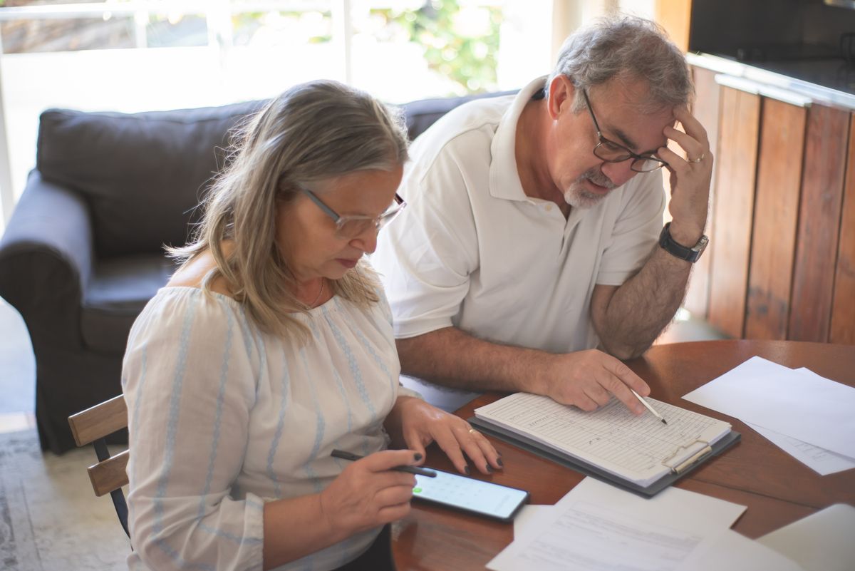 An older couple at a wooden table at home going through insurance paperwork with a pen and a phone calculator, the kind of side-by-side comparison of Medigap Plan G and Plan N premiums and copays that people in Delta, Cedaredge, Paonia and Hotchkiss work through